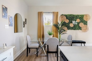 A modern dining area with a glass table, two chairs, a potted plant, and a wall-mounted decorative panel. - 3BR Gated Community Houston Home  + Parking (Houston)