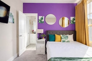 A bedroom with a purple wall, a bed with multiple pillows, a nightstand, and a mirror. - 3BR Gated Community Houston Home  + Parking (Houston)