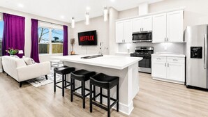A modern kitchen with white cabinets, a central island with bar stools, a microwave, and a refrigerator. - Designer 3BR w/ Fireplace, Yard & Free Parking (Houston)