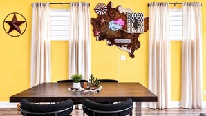 A dining area with a wooden table, two chairs, a wall-mounted decoration, and a window with curtains. - Gated Community Texas Style  3BR + Private Parking (Houston)