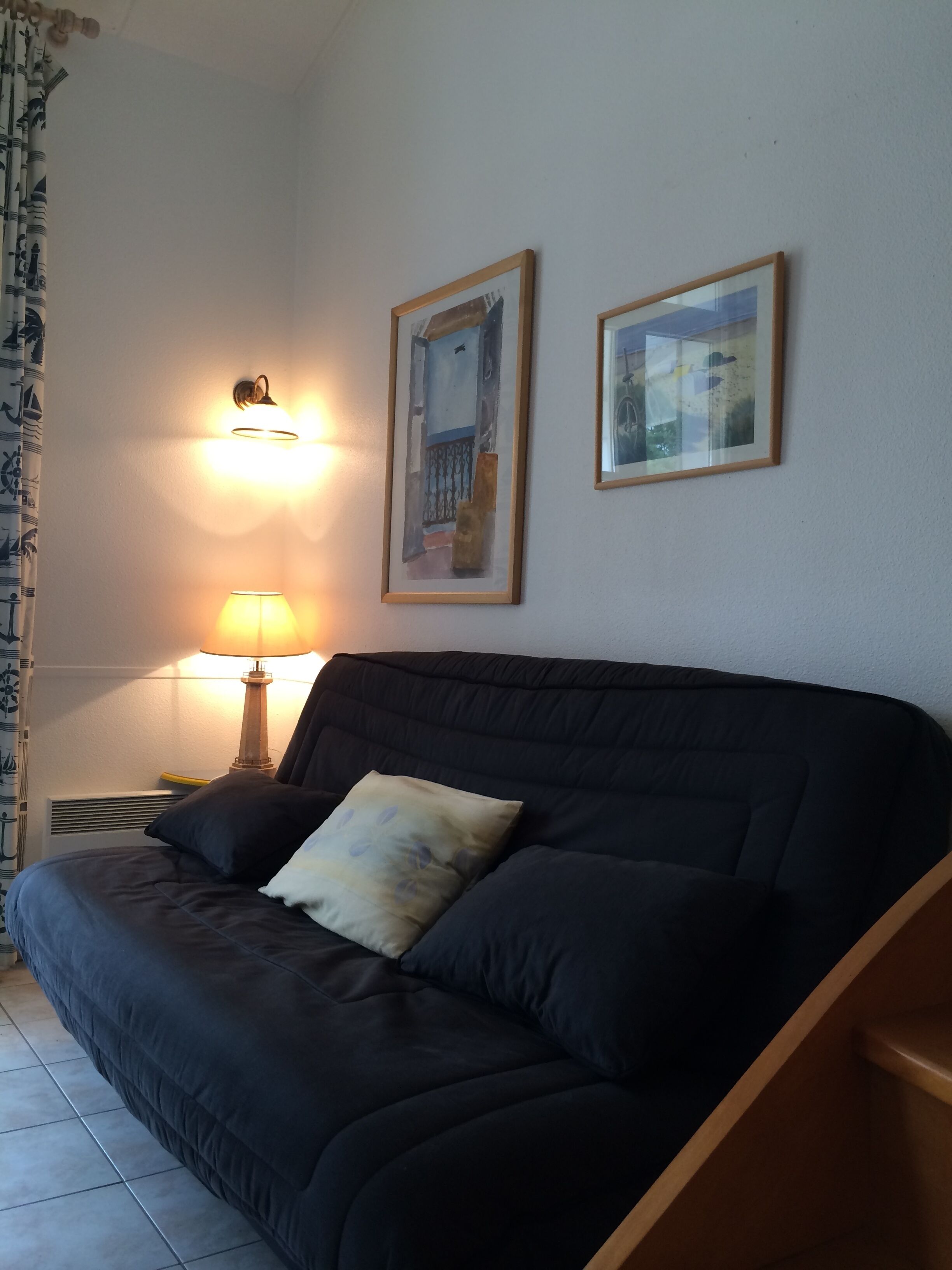 Un canapé de couleur sombre, agrémenté d’un seul coussin, une lampe fixée au mur et deux tableaux encadrés accrochés au mur.