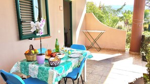 A dining table set outdoors with a view of the sea, a window with shutters, and a folding table.