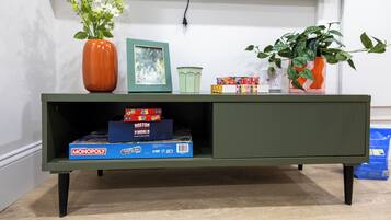 Een groene entertainmentkast met een televisie erboven, een vaas met witte bloemen, een ingelijst plaatje en een bloempot met plant.