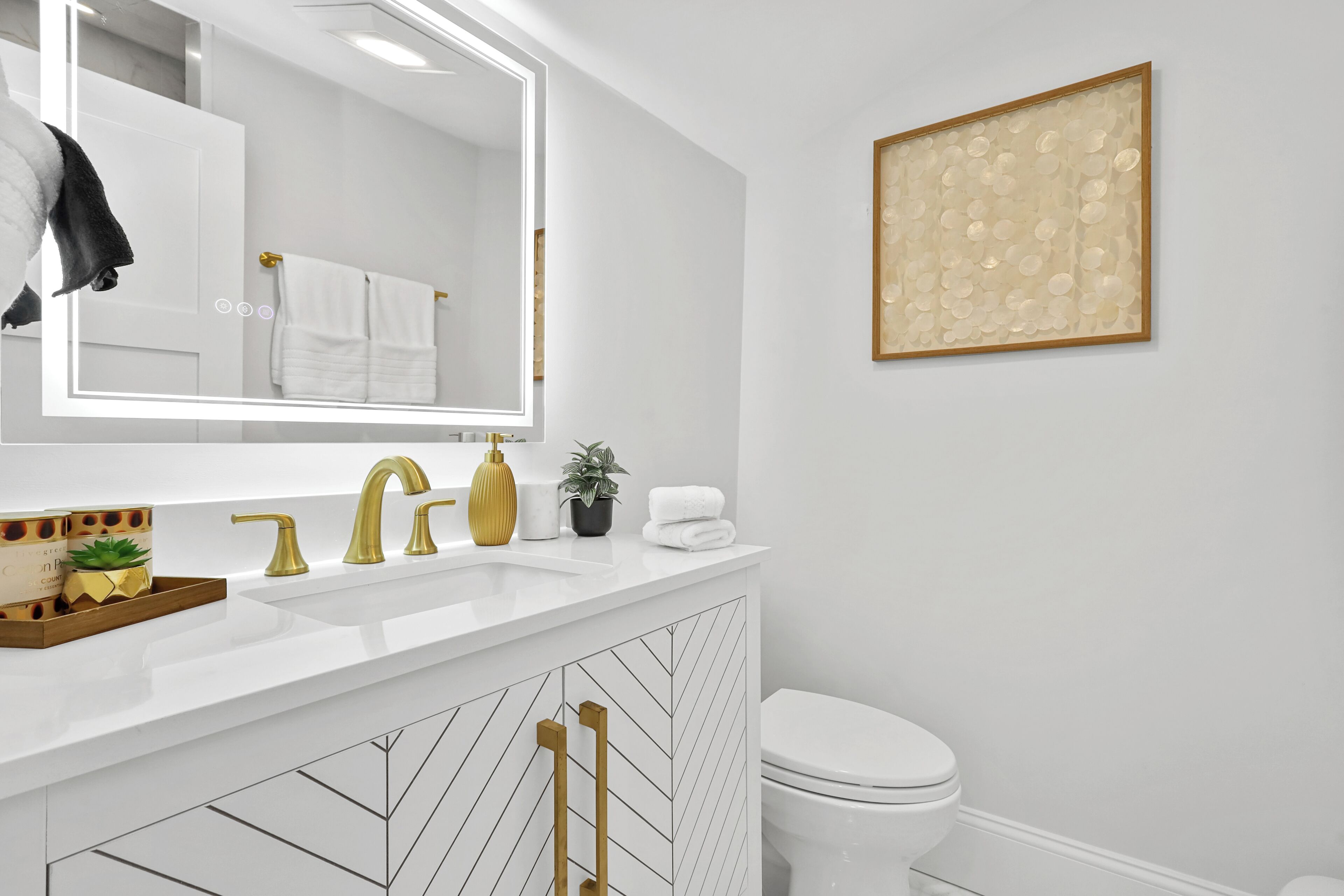 Ein modernes Badezimmer mit einem weißen Waschtisch, goldenen Armaturen, einem großen Spiegel und einem gerahmten Kunstwerk an der Wand.