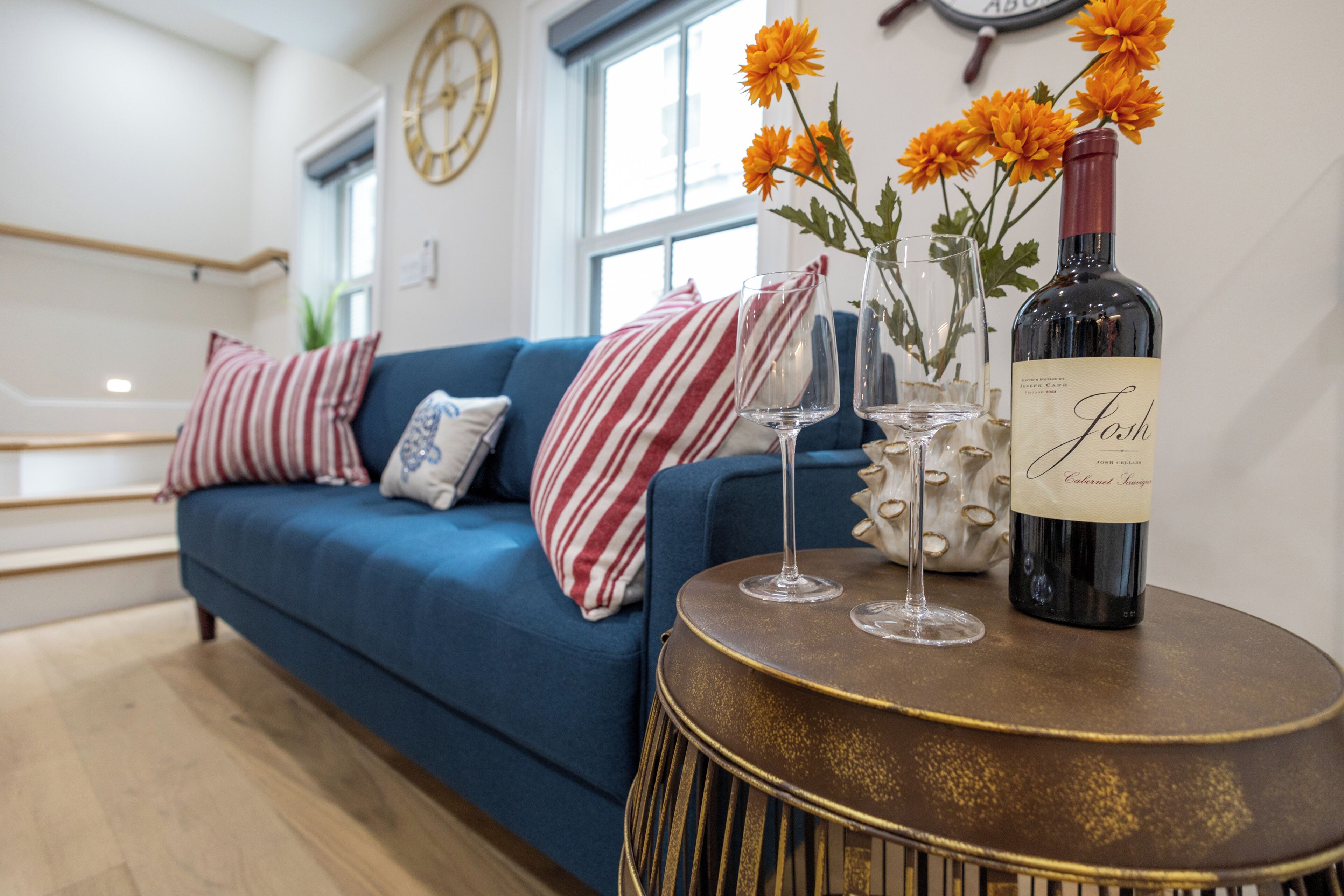 A living room with a blue sofa, striped pillows, a vase with orange flowers, two wine glasses, and a bottle of wine on a round table.