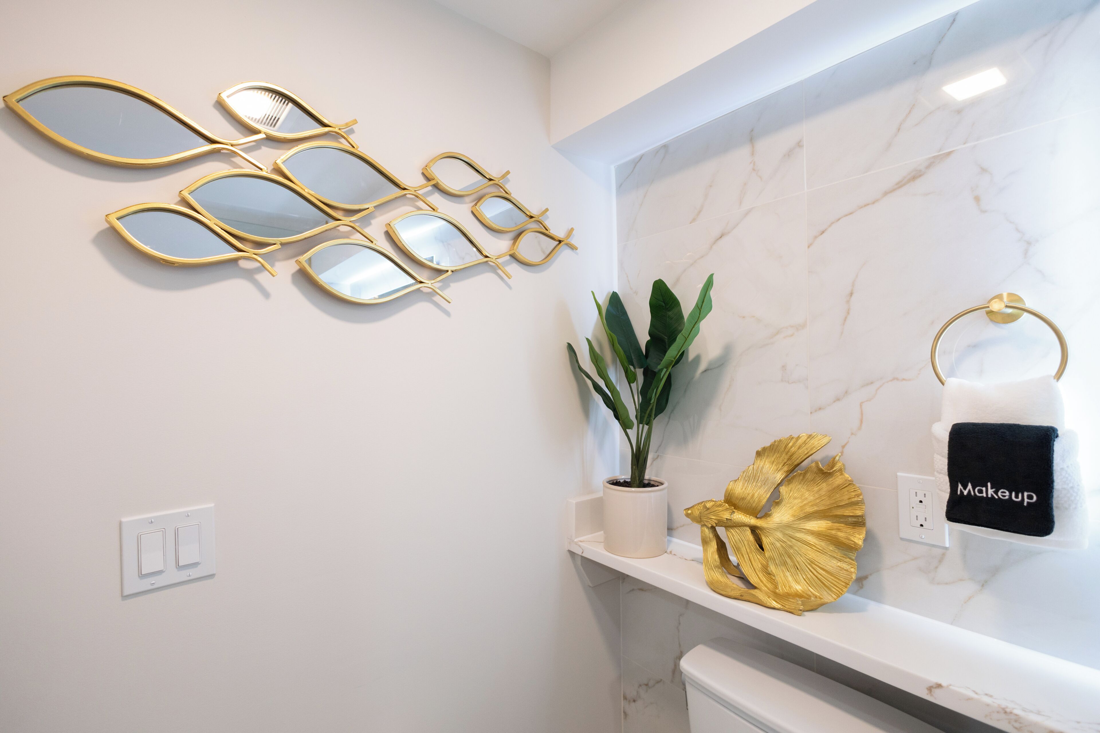Ein Badezimmer mit weißer Marmorwand, spiegeln in Goldrahmen, einer Topfpflanze, einem goldenen Dekorationsstück und einem Handtuchhalter mit einem Handtuch mit der Aufschrift 'Makeup'.