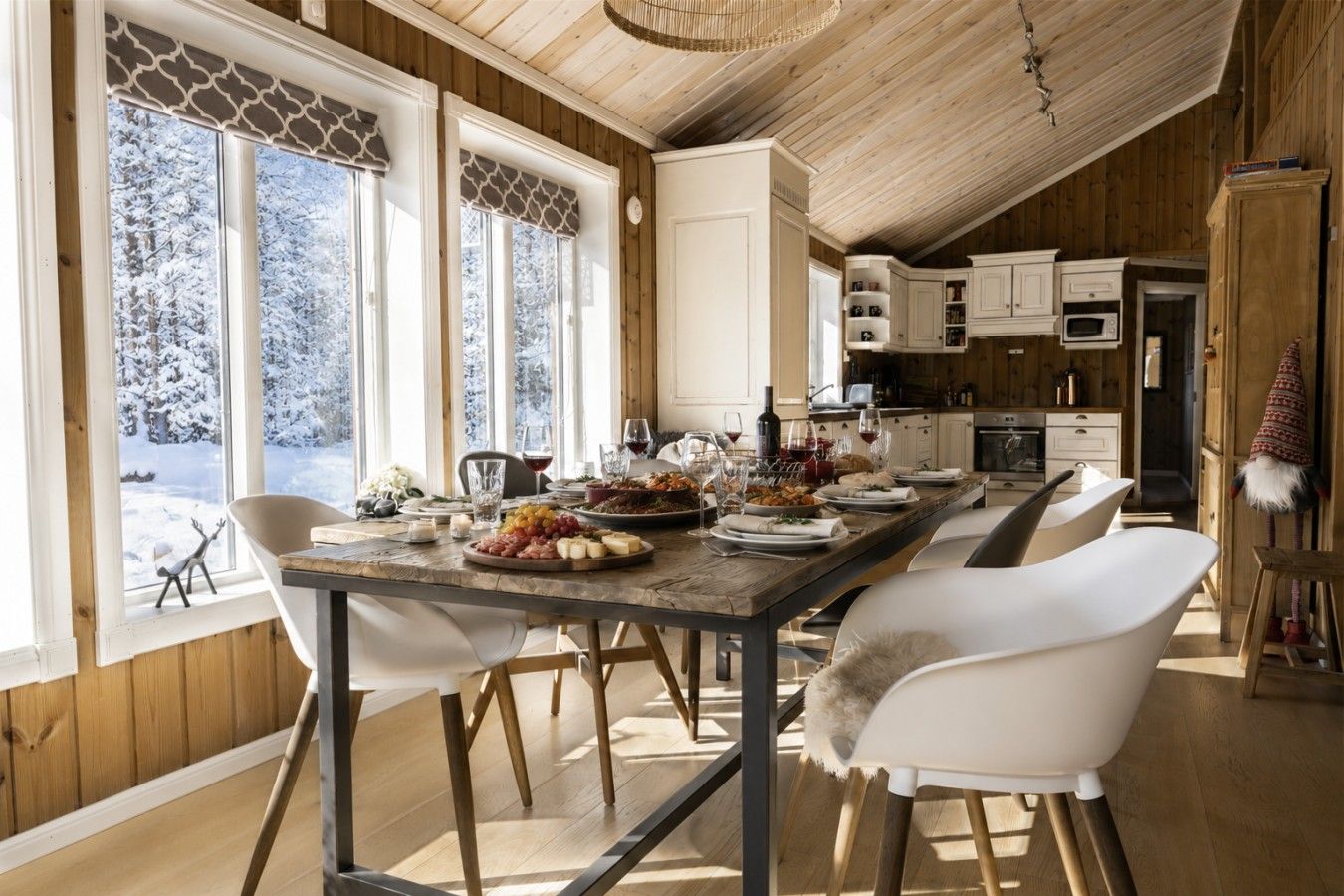 A dining area with a wooden table set for a meal, large windows with a view of snow-covered trees, and a kitchen in the background.