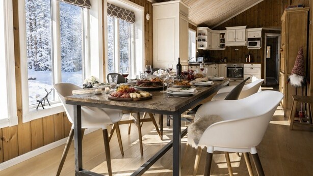 Een eethoek met een houten tafel, gedekt voor een maaltijd, omringd door witte stoelen, met uitzicht op besneeuwde bomen door grote ramen.