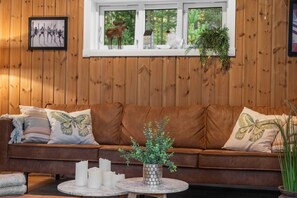 A cozy room with a brown leather sofa, a round marble table with candles, and a window with a view of trees. - The Cozy Family Mountain Retreat (Nybergsund)