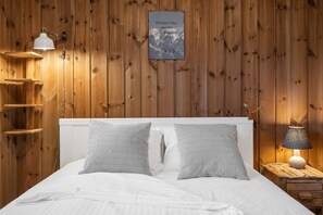 A wooden-paneled room with a bed, two patterned pillows, a wall-mounted lamp, a bedside table with a lamp, and a framed quote on the wall. - The Cozy Family Mountain Retreat (Nybergsund)