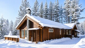 Een houten hutje met een stenen schoorsteen, omgeven door met sneeuw bedekte bomen.