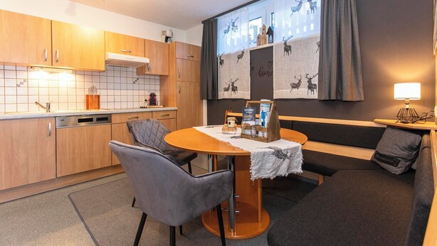 A modern kitchen with wooden cabinets, a dining table with chairs, and a cozy seating area with a sofa and a lamp. - Gruber's Gästehaus - Ferienwohnungen (Viehhofen)