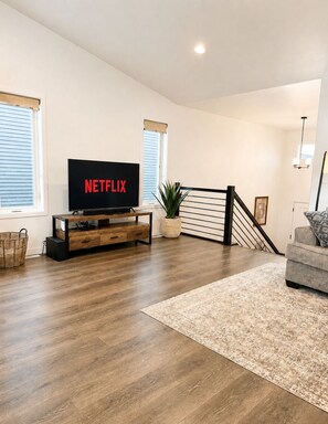 Ein modernes Wohnzimmer mit einem Flachbildfernseher, der 'NETFLIX' anzeigt, einer Holz-Unterhaltungseinheit, einer Treppe und einem gemusterten Teppich.