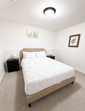 A bedroom with a bed, two bedside tables, a wall-mounted light fixture, and two framed artworks.