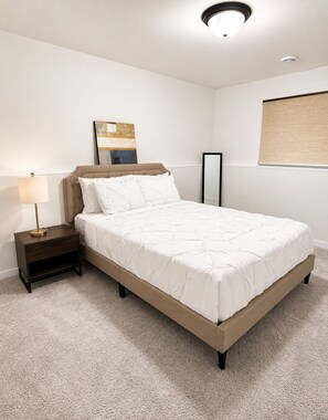 Um quarto com cama, mesa de cabeceira, abajur e uma obra de arte na parede.