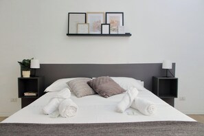 Ein ordentlich bezogenes Bett mit aufgerollten Handtüchern, ein grauer Kopfteil und ein wandmontiertes Regal mit gerahmten Bildern und einer kleinen Pflanze.