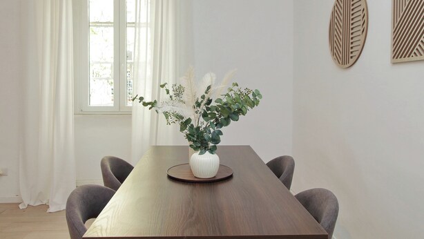 A dining table with a white vase of greenery, two wall art pieces, and a window with white curtains. - Amiterno Elegance - by Domus & Dimore (Rome)