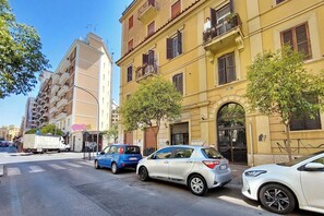 Straßenansicht mit parkenden Autos, einem Gebäude mit gelber Fassade und einem baumbewachsenen Bürgersteig.