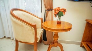Una habitación con una silla de madera, una pequeña mesa redonda de madera con un florero y una ventana con cortinas.