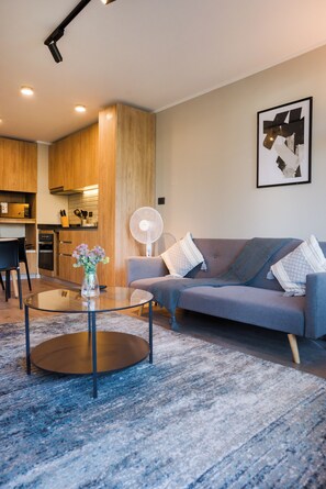 Ein modernes Wohnzimmer mit einem Glastisch, einem grauen Sofa und einer Küche im Hintergrund.