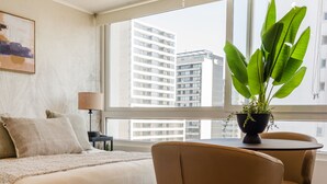 Et moderne hotelværelse med en stor seng, et skrivebord med stol, en potteplante og udsigt til byens bygninger gennem vinduet.