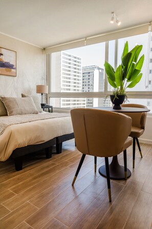 Ein modernes Hotelzimmer mit einem großen Bett, einem Schreibtisch mit Stuhl, einer Zimmerpflanze und Blick auf Stadtgebäude durch das Fenster.