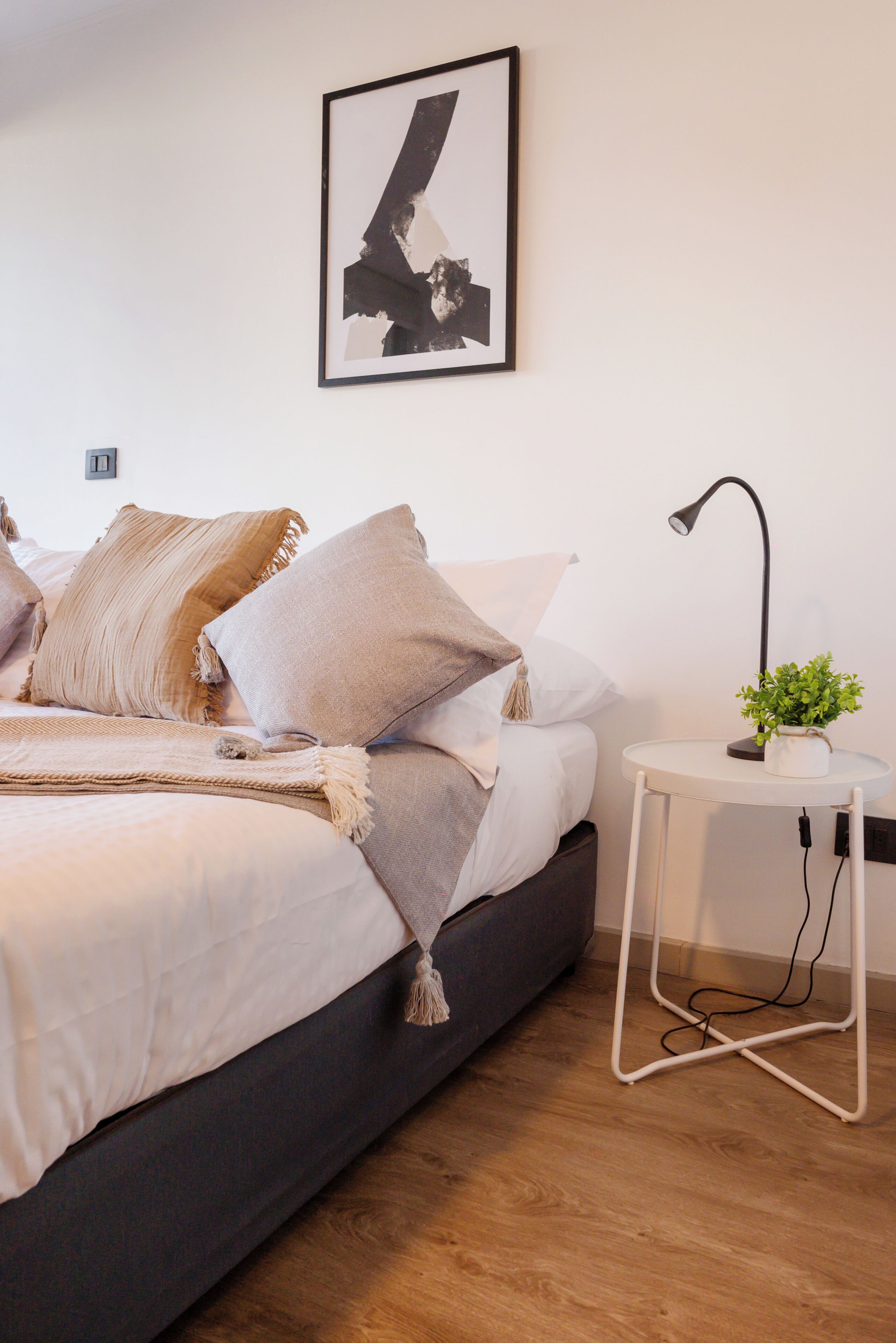 A modern bedroom with a bed, pillows, a bedside table with a lamp, and a framed picture on the wall.