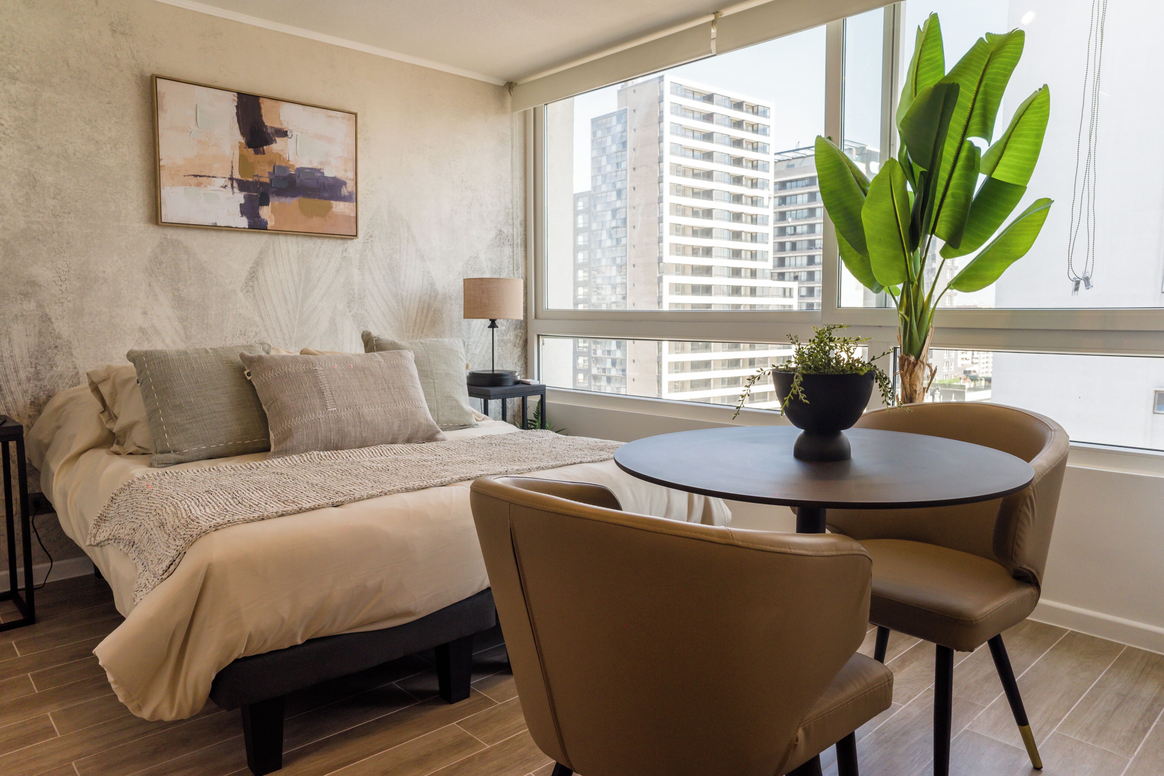 A modern hotel room with a bed, a round table, a chair, a lamp, and a large window with a view of buildings.