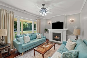 A living room with a sofa, a coffee table, a fireplace, a television, and a ceiling fan. - La Dolce Vita | Walk to Downtown NOTL (10 minutes) (Niagara-on-the-Lake)