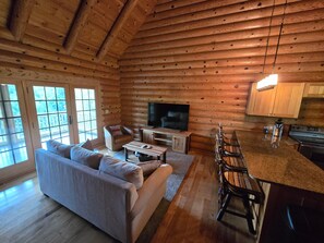 A log cabin interior with a sofa, TV, dining table, chairs, and a kitchen area.