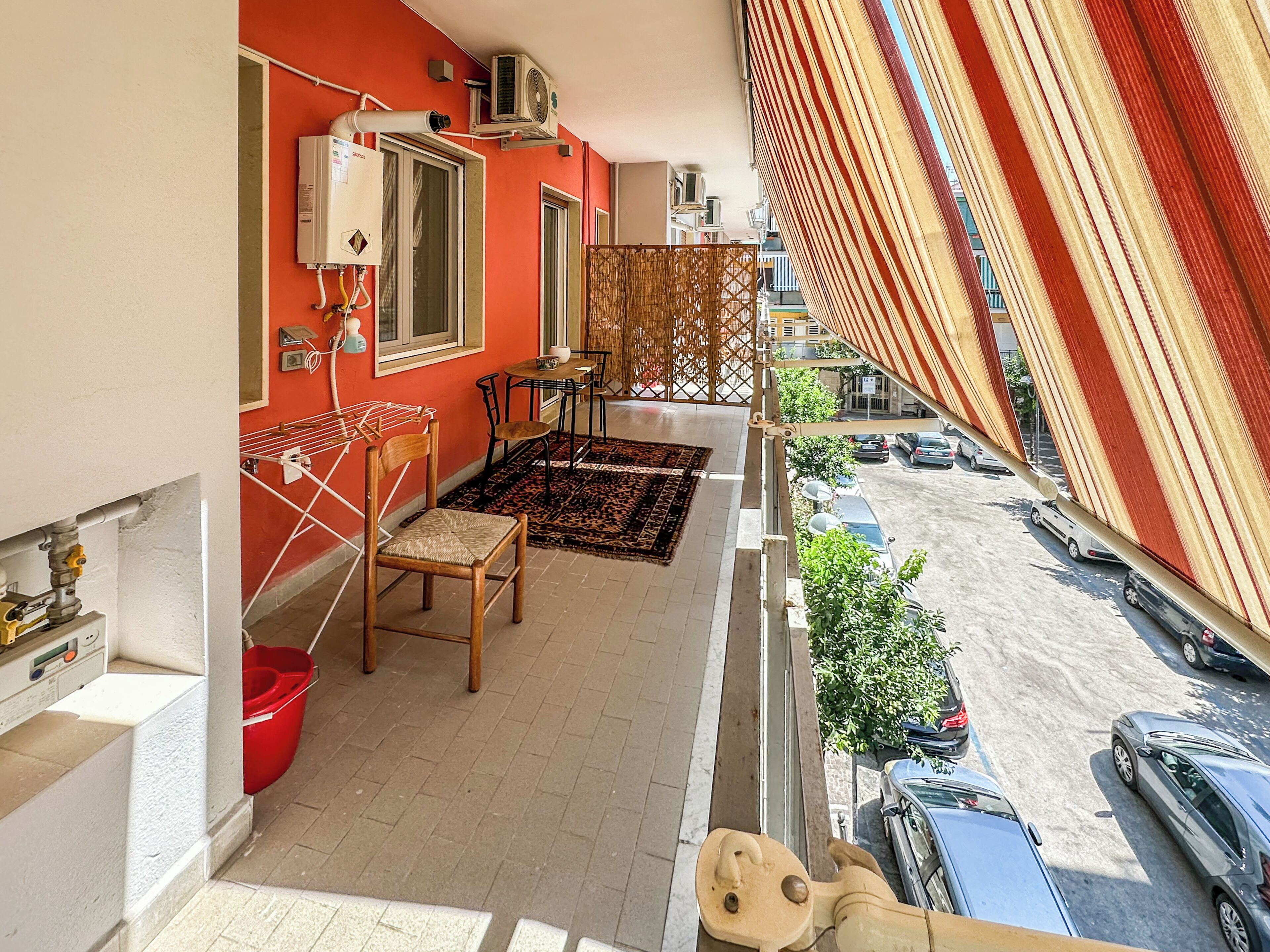 Un balcon avec un mur rouge, un lave-linge, une chaise, une table et une vue sur les voitures et les immeubles garés.