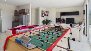 Una sala de estar moderna con una mesa de fútbol, un comedor con mantel rojo, una cocina y un televisor al fondo.