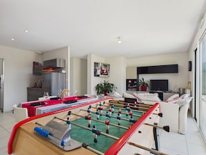 A modern living room with a foosball table, a dining area with a red tablecloth, a kitchen, and a TV in the background. - House for 4, Jacuzzi & Garden, Beach 200 m Away (Plouguerneau)