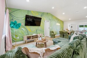 A living room with a green patterned wall, a flat-screen TV, a sofa, a coffee table, and a dining area with chairs. - Pickleball, Hot Tub, Pool, Mini Golf, Arcades, Billiards, and More! (St. Petersburg)