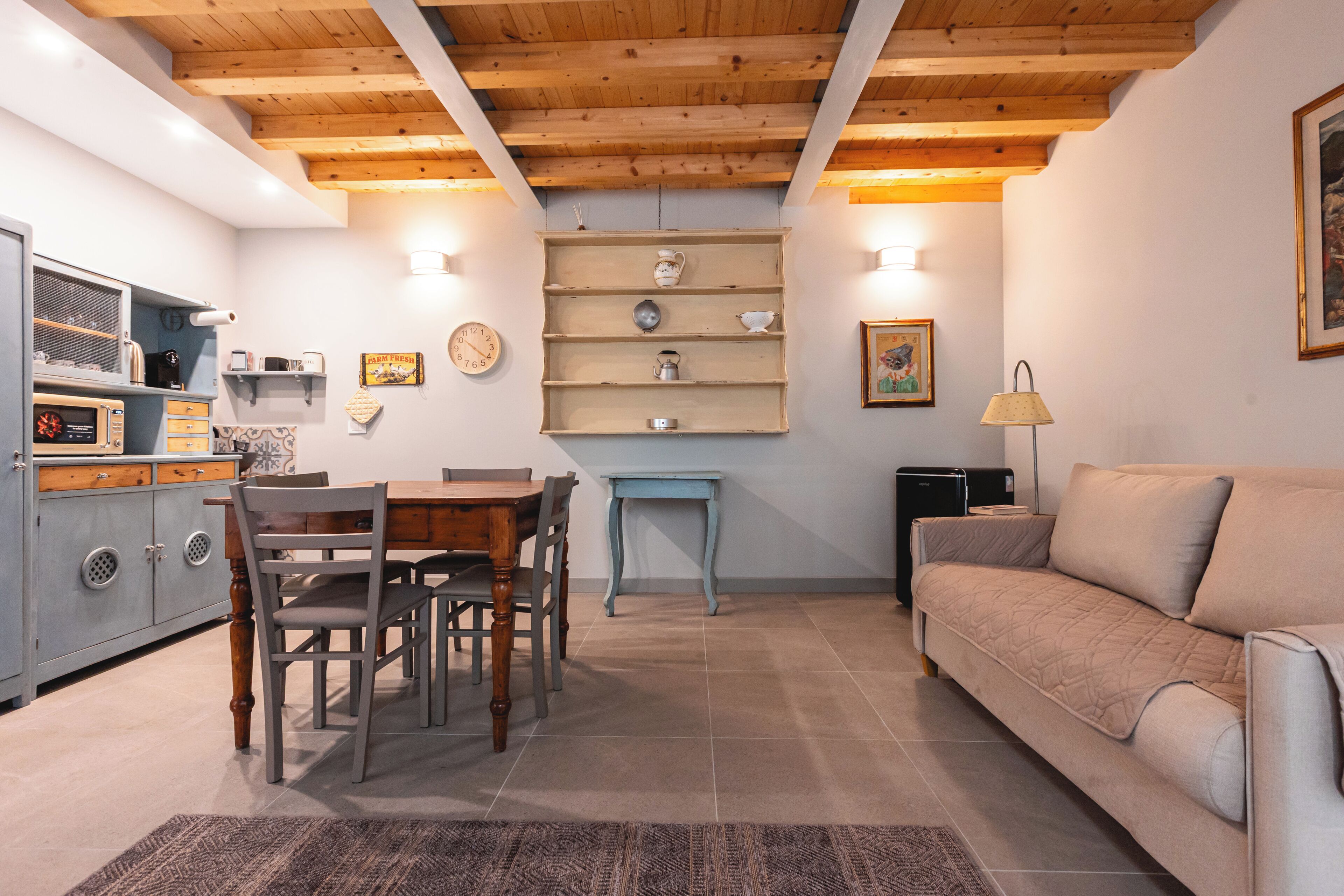 Un salon chaleureux avec un plafond en bois, un coin repas avec une table et des chaises, un canapé et un espace cuisine équipé d’appareils électroménagers et d’étagères.