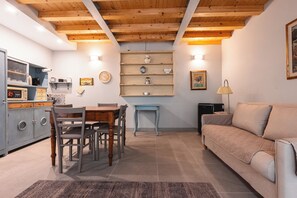 Una sala de estar acogedora con techo de madera, comedor con mesa y sillas, sofá y zona de cocina con electrodomésticos y estanterías.