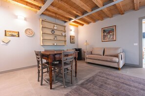 A dining area with a wooden table and chairs, a sofa, a clock, and a shelf with kitchenware.