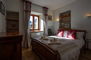 Ein Schlafzimmer mit einem hölzernen Bett, einem Fenster mit Vorhängen, einem Nachttisch und einem Regal mit Dekorationen.