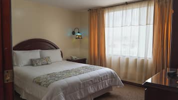 Quarto de hotel com cama, mesa de cabeceira, luminária e janela com cortinas.