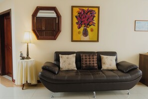 A living room with a dark leather sofa, a round mirror, a table with a lamp, and a framed painting on the wall.