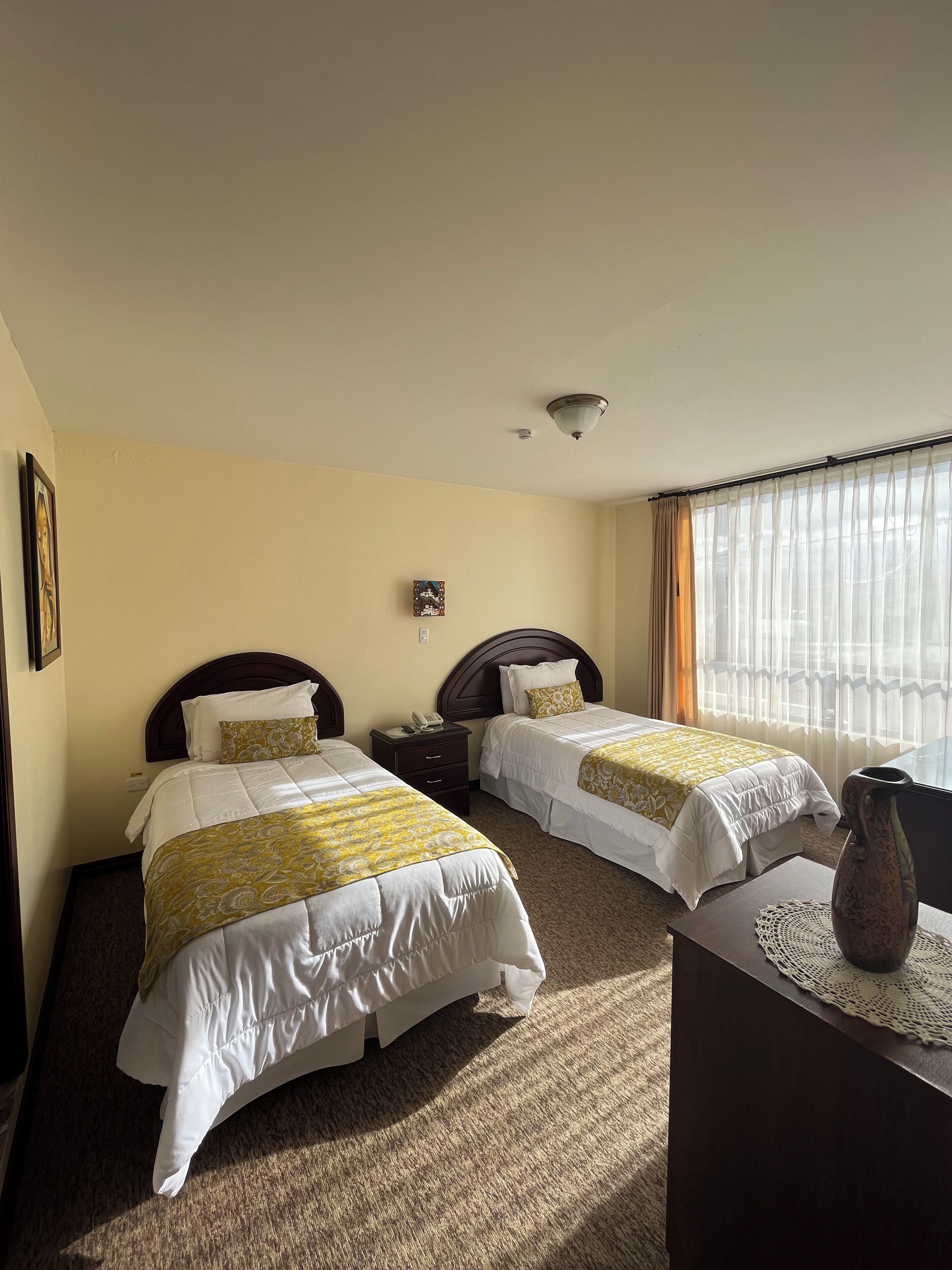 A hotel room with two beds, a bedside table, a lamp, and a window with curtains.