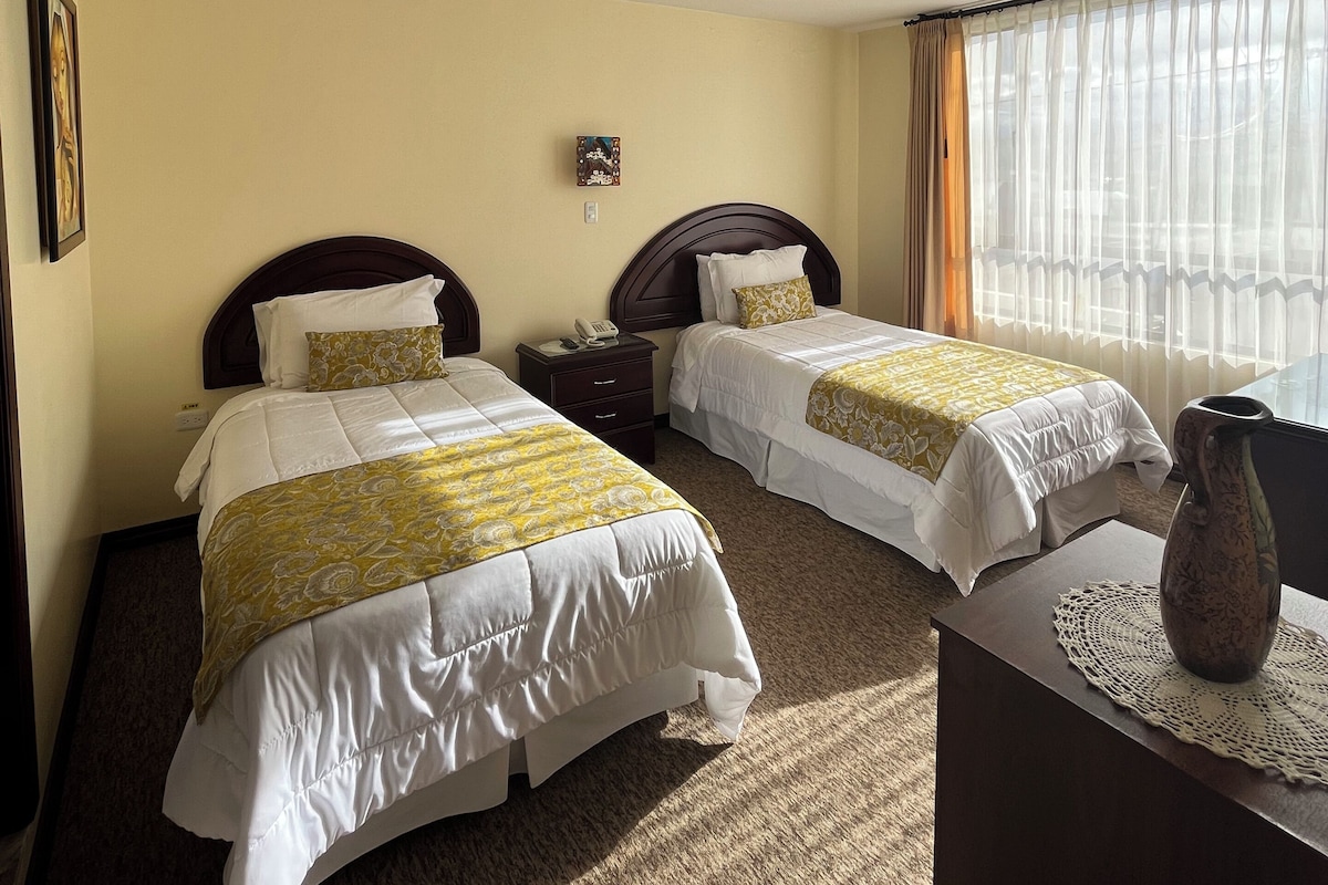 A hotel room with two beds, a bedside table, a lamp, and a window with curtains.