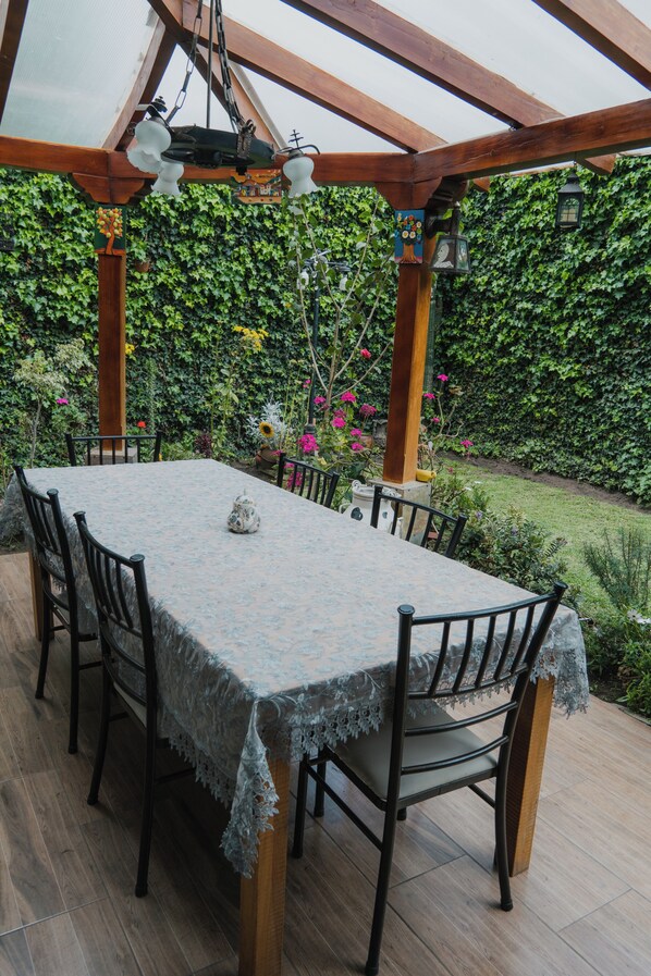 A covered patio with a table and chairs, surrounded by greenery and a garden.