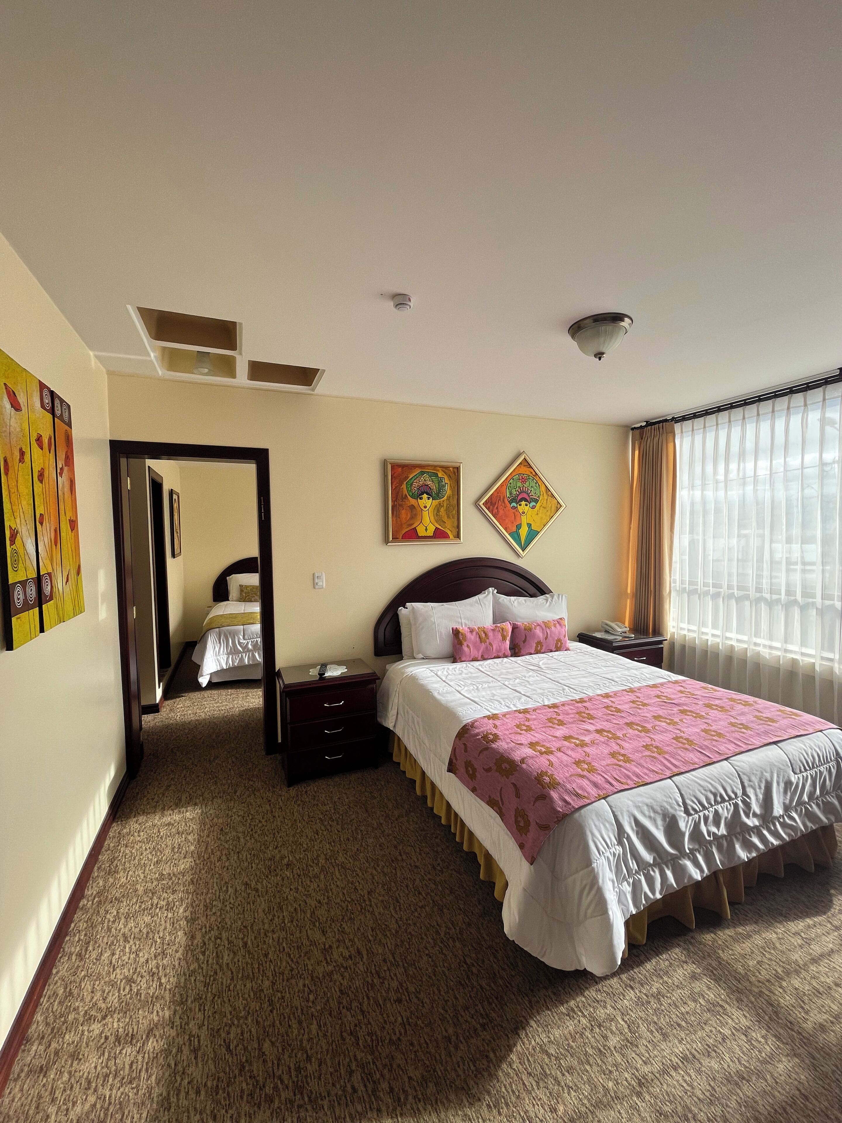 Een hotelkamer met een groot bed, een nachtkastje, een wanddecoratie, een deur naar een andere kamer en een raam met gordijnen.