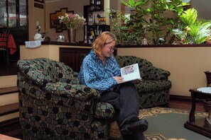 A person seated on a patterned sofa in a room with a wooden floor, a small table, and various decorative items including a vase with flowers.