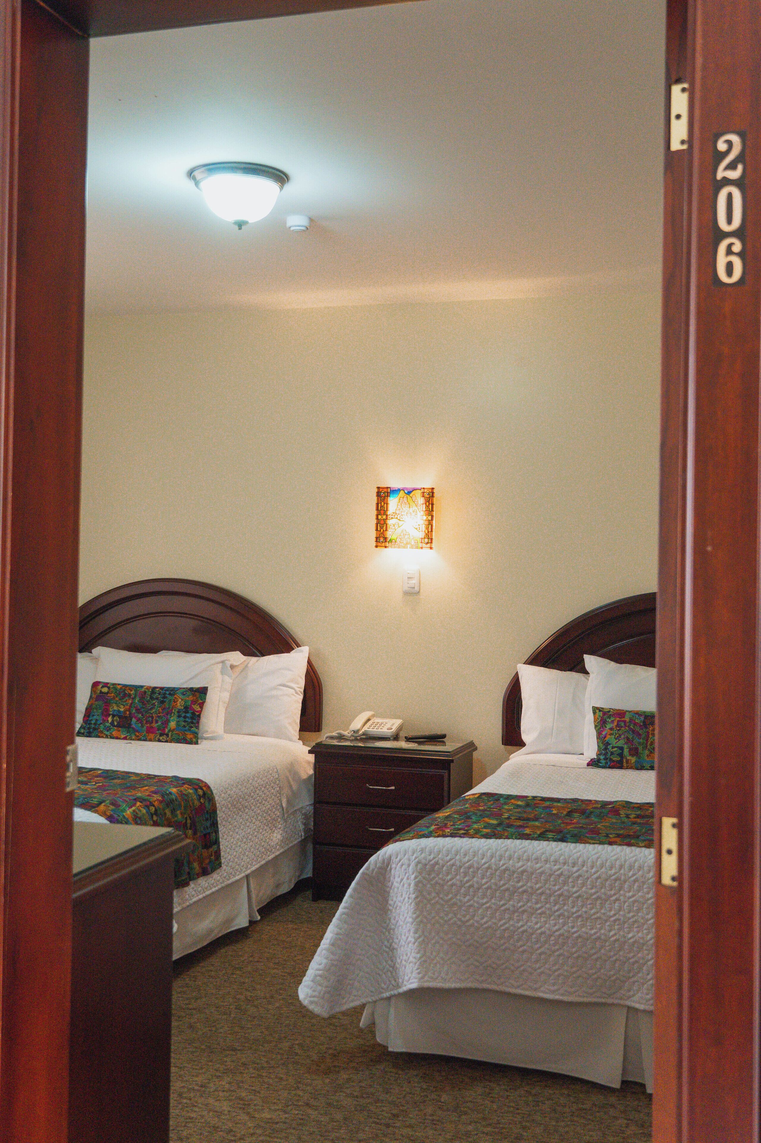 A hotel room with two beds, a nightstand with a phone, and a wall-mounted light fixture.