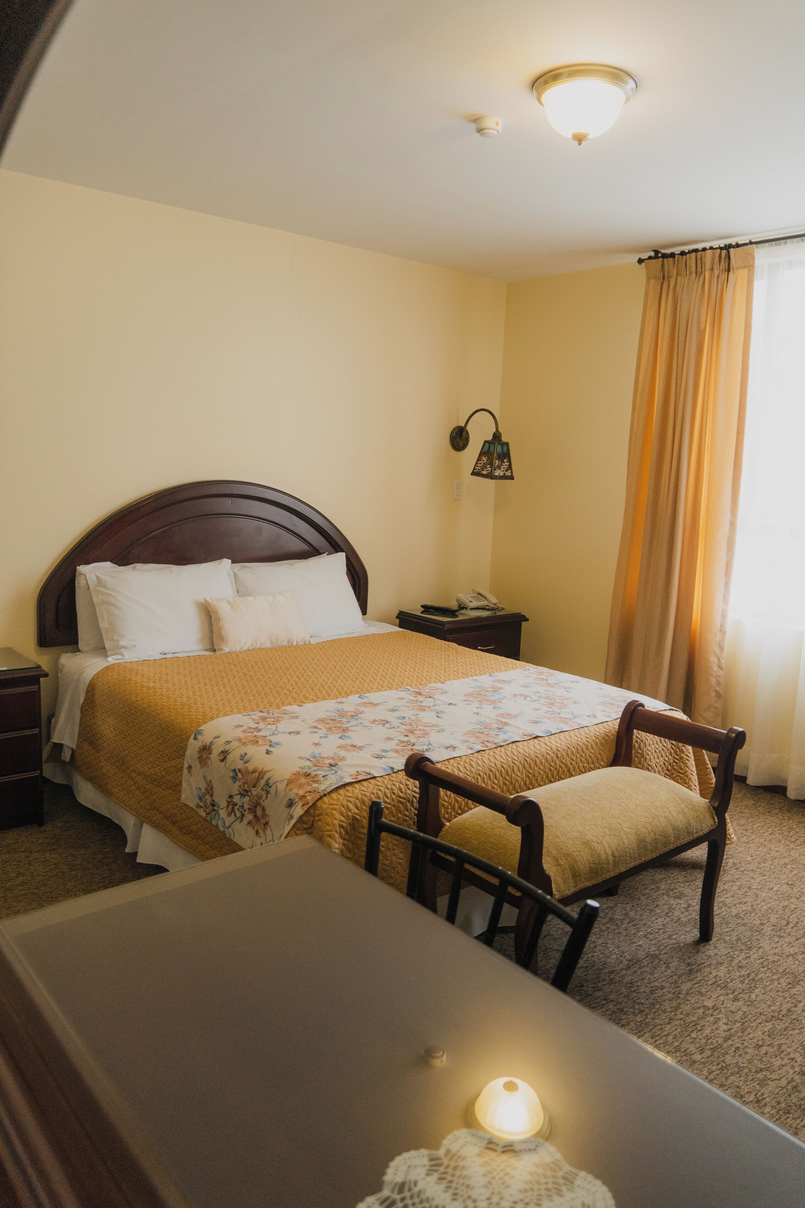 A hotel room with a bed, bedside table, lamp, and a view of the outside through a window with curtains.