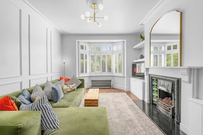 A modern living room with a green sofa, a fireplace, and a bay window with greenery. - Knutsford Home | Sun Trap Garden | Sleeps 8 (Cheshire East)