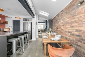 A modern kitchen with a central island, bar stools, and a dining area set for a meal. - Knutsford Home | Sun Trap Garden | Sleeps 8 (Cheshire East)