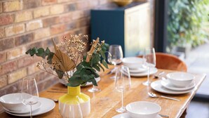 Una mesa de comedor dispuesta con platos, fuentes y copas de vino blancos, con un jarrón amarillo que contiene flores secas y hojas verdes.